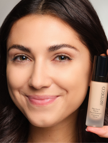 Model mit dezentem Makeup Look hält eine Foundation neben dem Gesicht
