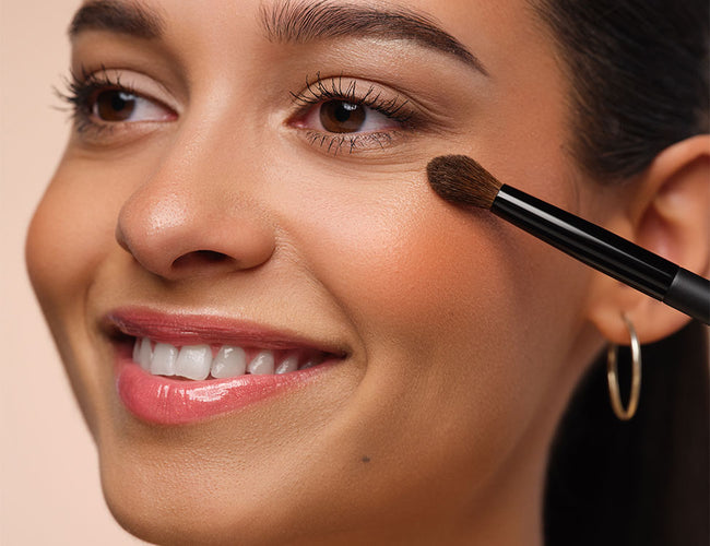 Highlighter wird mit einem kleinen, fluffigen Pinsel auf den höchsten Punkt des Wangenknochens vom Model aufgetragen