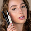 Model mit getuschten Wimpern hält zwei verschiedene Mascaras neben das Gesicht.