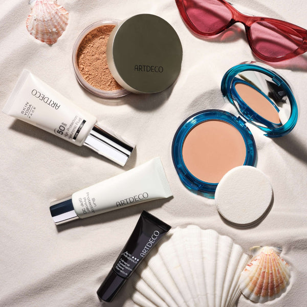 Artdeco Produkte mit Sonnenschutz und andere Sommerartikel, wie Sonnenbrille und Muscheln platziert auf sandigem Hintergrund. 