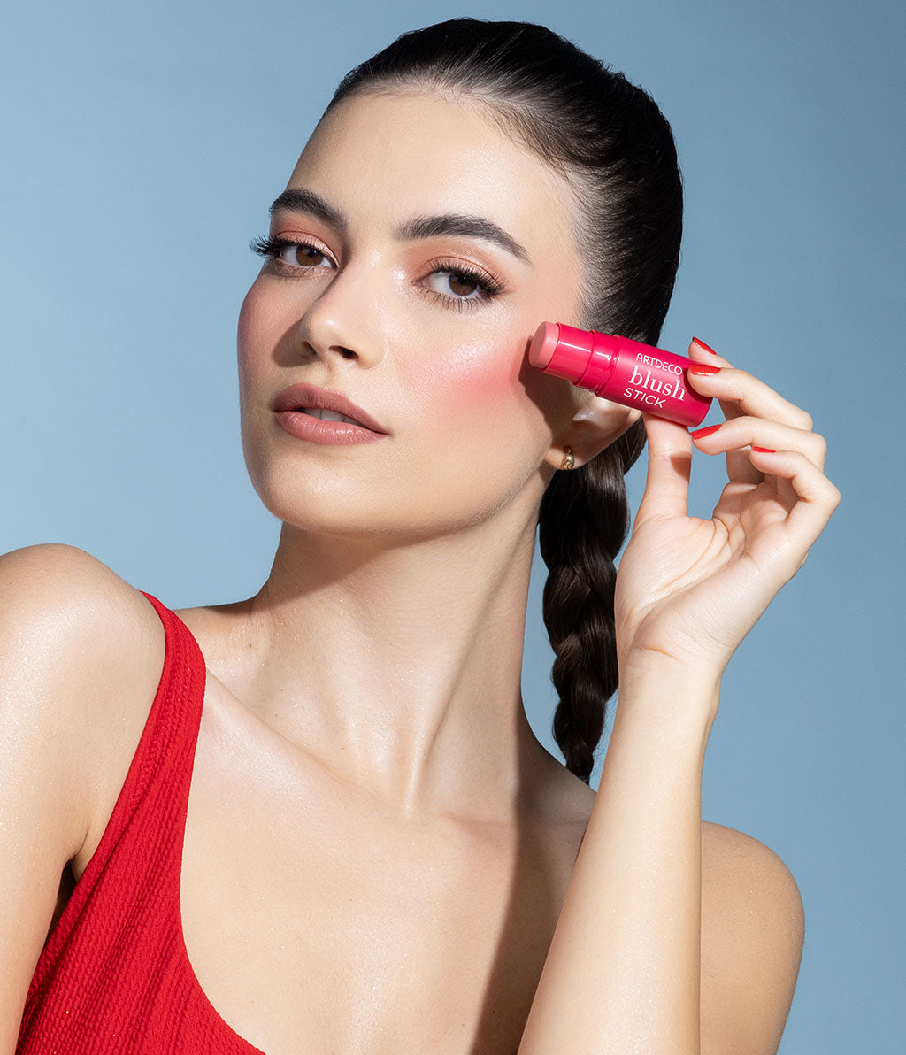 Eine Frau mit einem natürlichen Make-up hält einen Blush Stick an ihre Wange und blickt sanft in die Kamera. Sie trägt ein rotes Oberteil und hat einen geflochtenen Zopf.