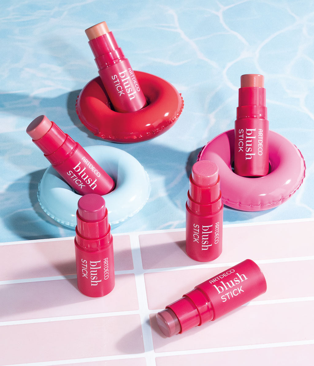 Sechs Blush-Sticks in verschiedenen Farben sind auf bunten Schwimmringen angeordnet, die auf einer Wasseroberfläche schwimmen. Die Produkte strahlen Frische und Sommerlichkeit aus.