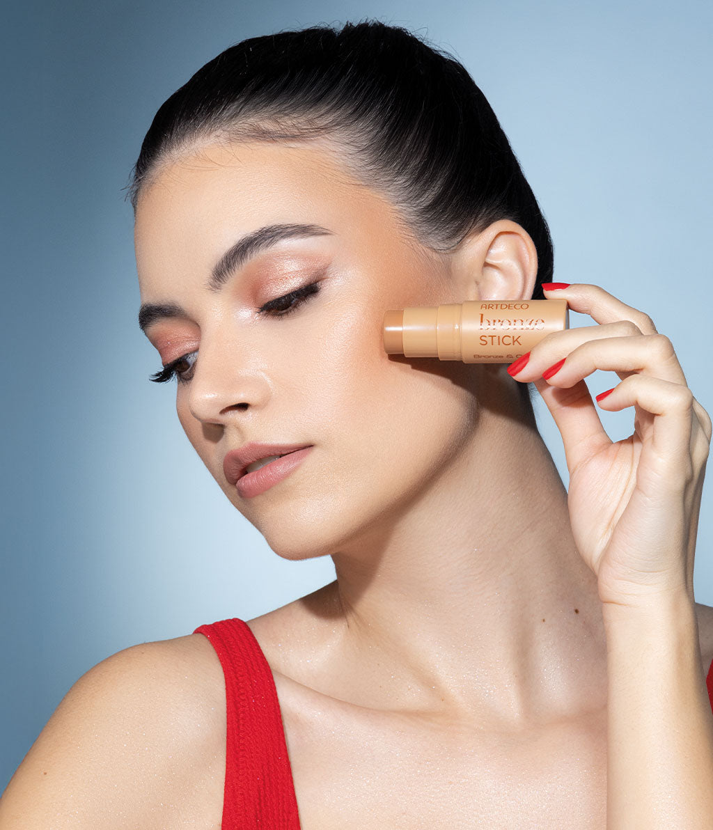 Eine Frau mit einem sommerlichen Make-up hält einen Bronze Stick an ihre Wange und schaut zur Seite. Sie trägt ein rotes Oberteil und hat einen streng geflochtenen Zopf.