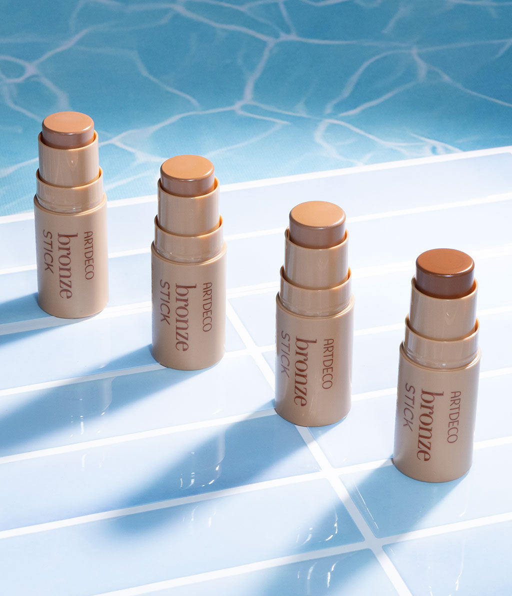 Vier Bronze Sticks stehen in einer Reihe auf Pool-Fliesen, während sie auf einer Wasseroberfläche reflektiert werden. Die Produkte strahlen Sommerlichkeit aus.