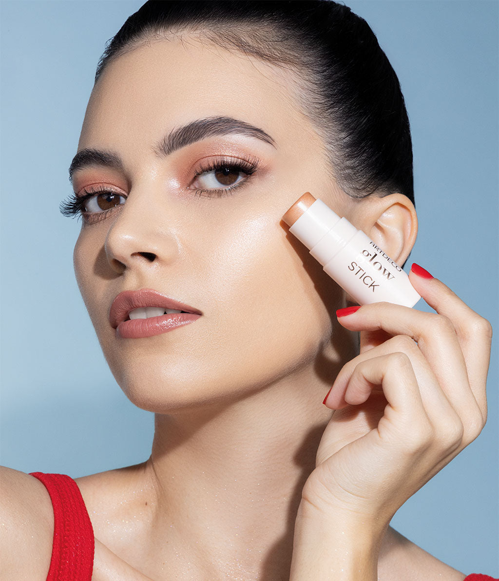 Eine Frau mit schimmerndem Make-up trägt mit einem ARTDECO Glow Stick Highlighter auf ihre Wangenknochen auf. Ihr Look ist sommerlich-frisch mit glänzender Haut und dezentem Lidschatten.