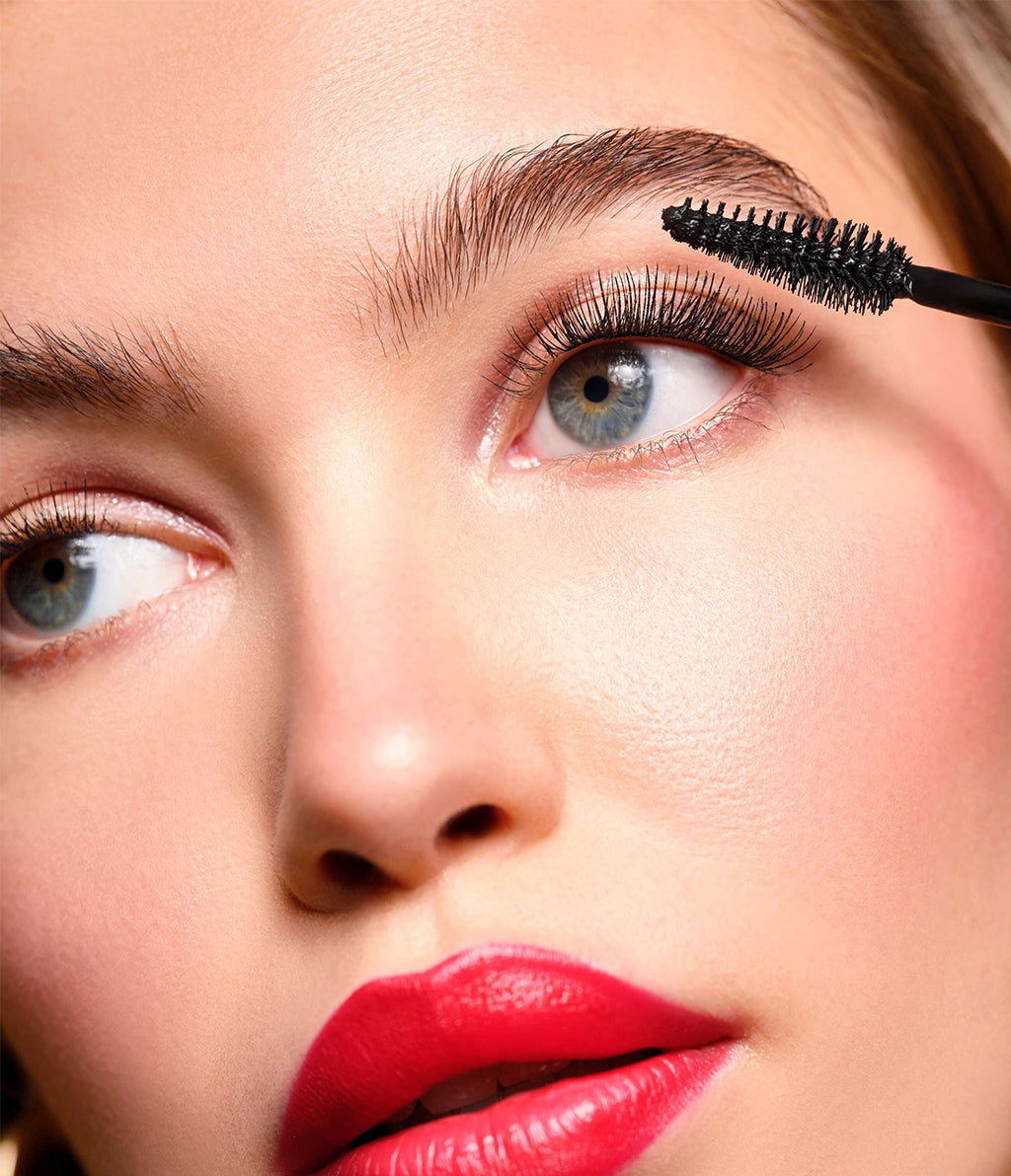 Eine junge Frau mit blonden Haaren, natürlichem Make-up und ausdrucksstarken roten Lippen trägt die Angel Eyes Mascara im limitierten goldenen Design auf; das Bürstchen ist dabei gut zu erkennen.