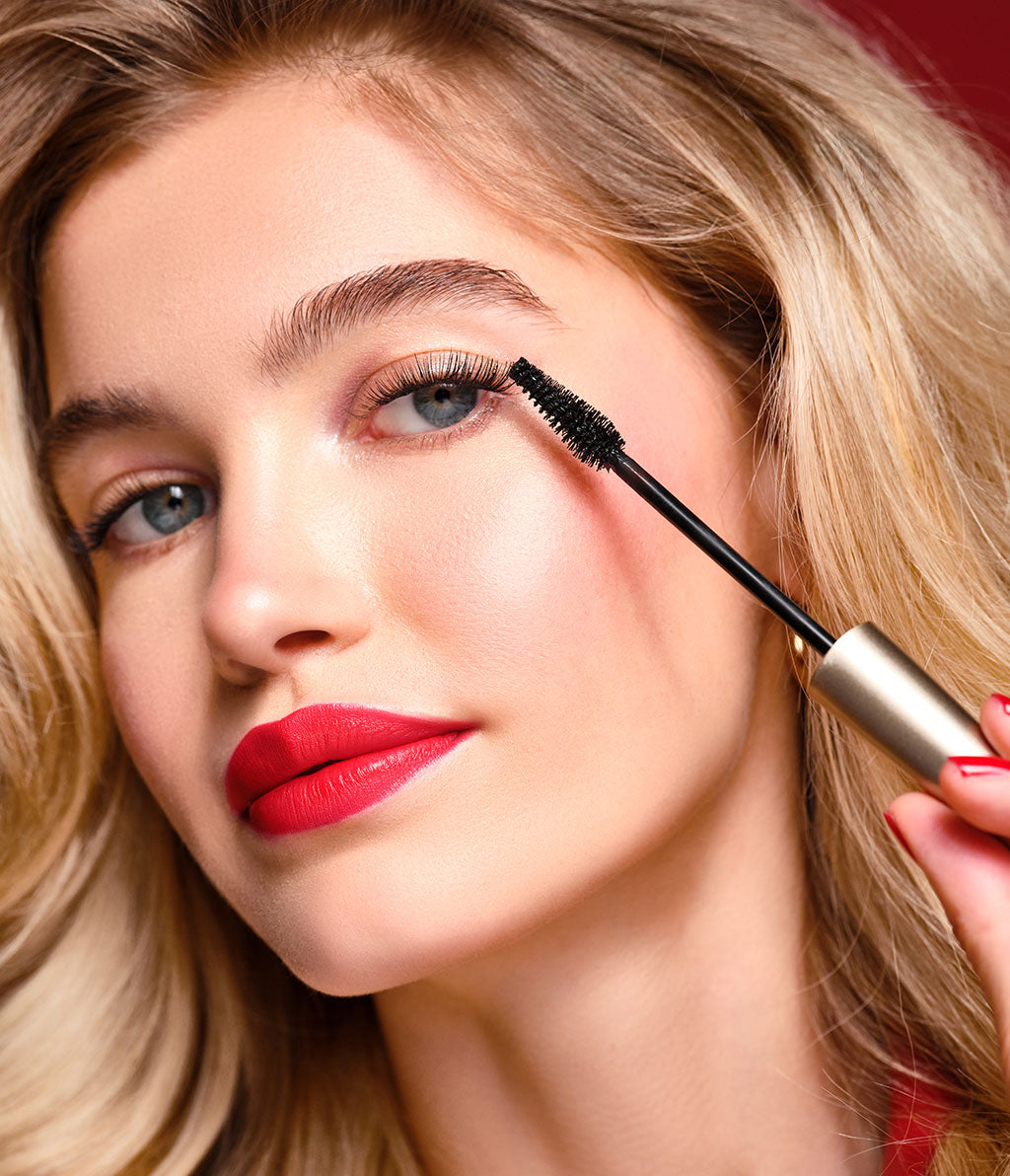 Eine junge Frau mit blonden Haaren, natürlichem Make-up und ausdrucksstarken roten Lippen trägt die Angel Eyes Mascara im limitierten goldenen Design auf; das Bürstchen ist dabei gut zu erkennen.