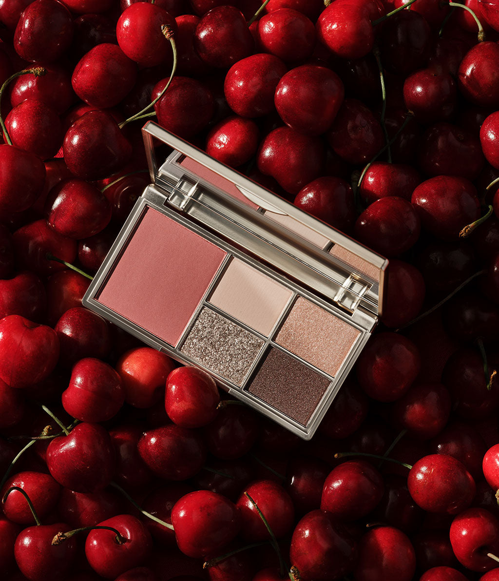 Geöffnete Illusion Palette for Cheeks & Eyes mit vier Lidschatten in Nude- und Champagner-Tönen sowie einem Blush im limitierten goldenen Design, liegend auf einem Untergrund aus Kirschen.