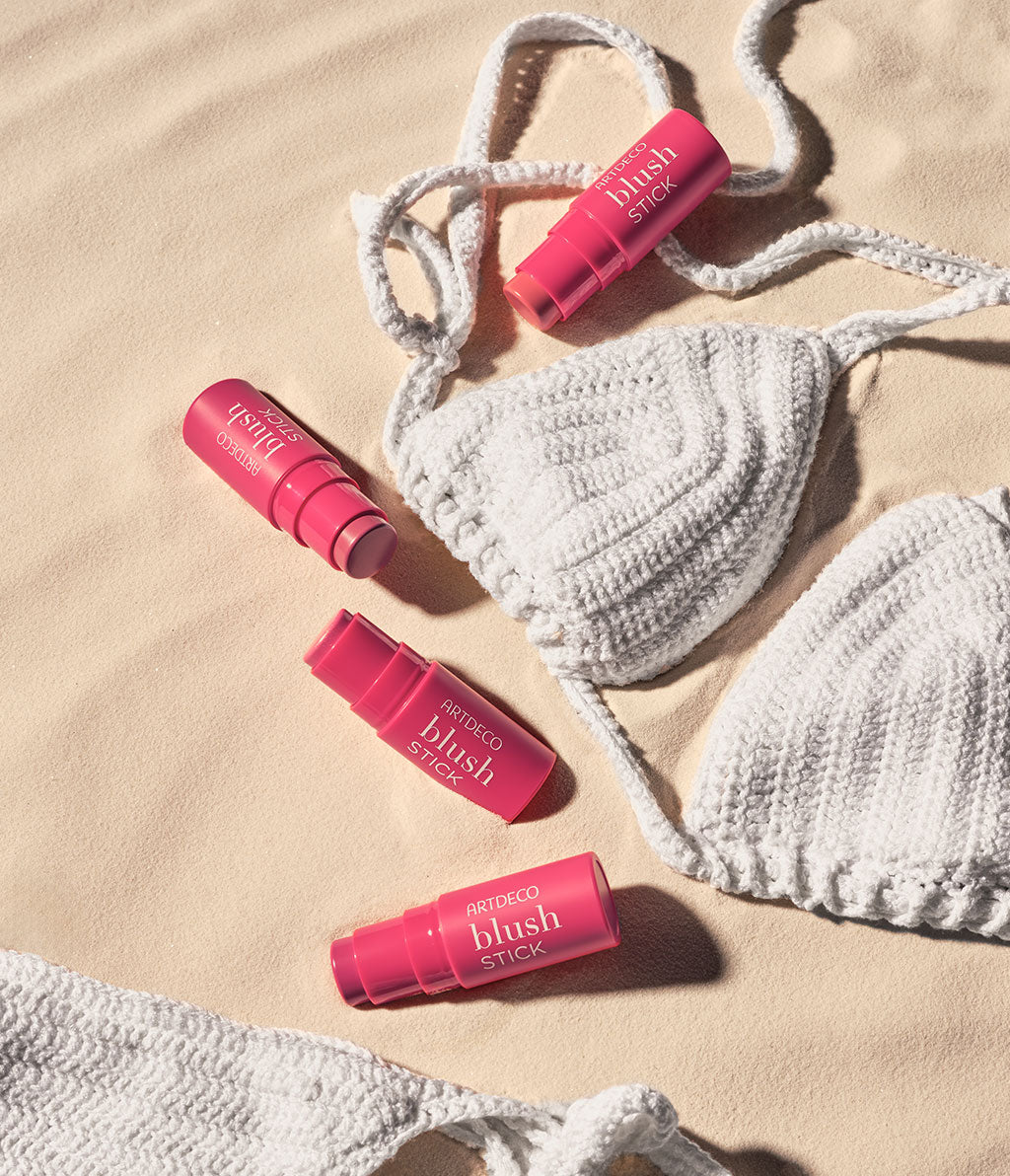 Mehrere Blush Sticks von ARTDECO liegen auf einem sandfarbenen Untergrund, umgeben von einem weißen, gestrickten Bikini.