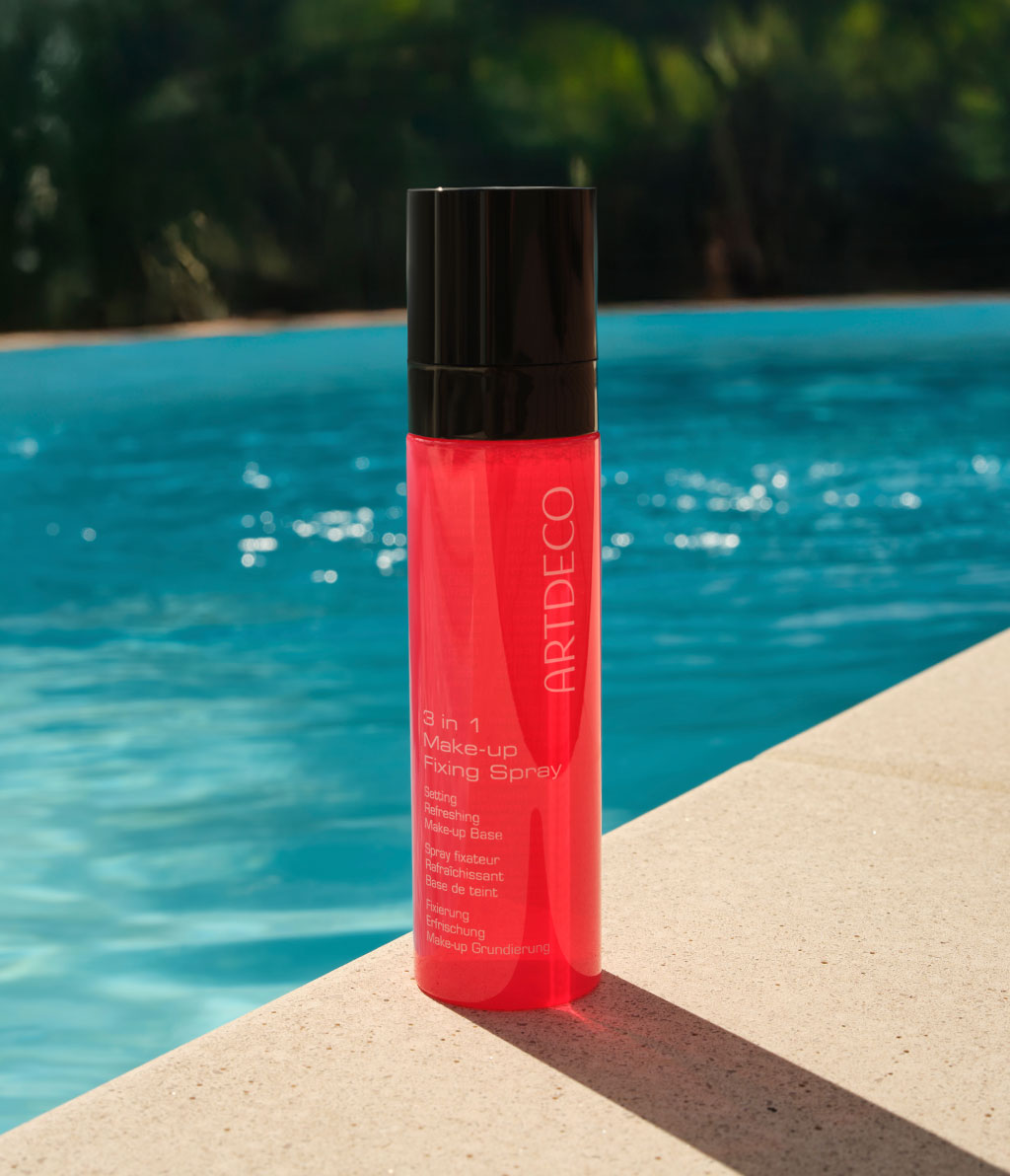 3 in 1 Fixing Spray in der roten Flasche steht am Rand einem Pools. Das Wasser funkelt im Hintergrund.