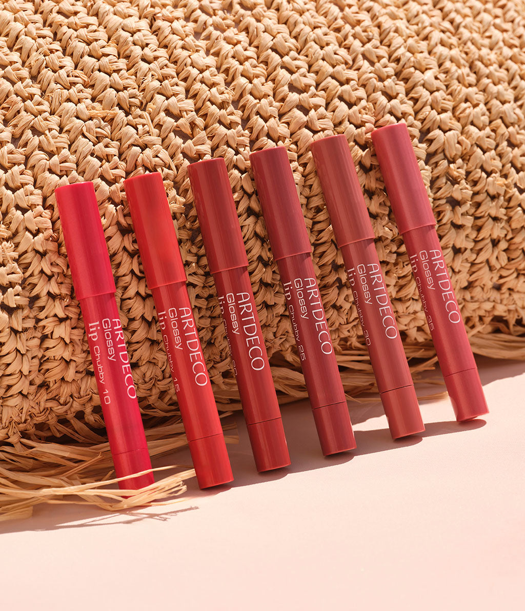 Alle sechs Farbe des Glossy Lip Chubby stehen gelehnt an einer Tasche aus Bast