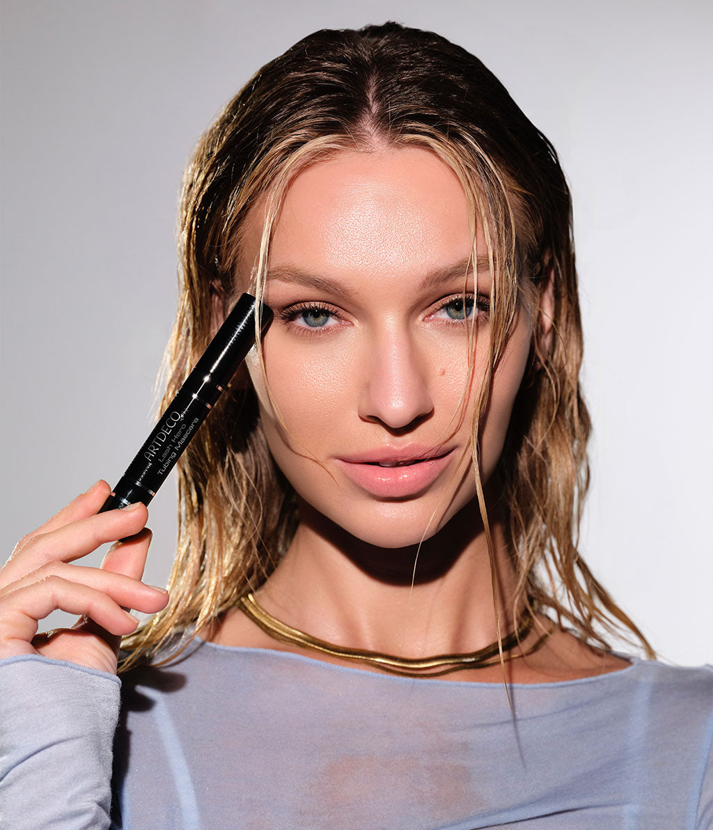 Model mit leicht angefeuchteten Haaren und Klamotten, hält die Lash Hero Tubing Mascara.