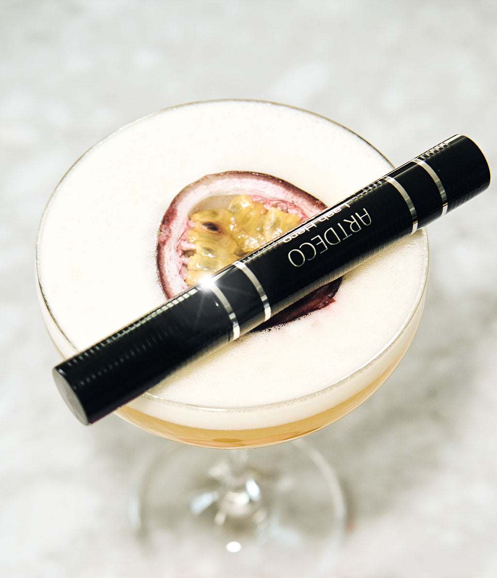 Geschlossene Lash Hero Tubing Mascara liegt auf einem Glas mit einem alkoholischen Getränk mit einer Passionsfrucht.