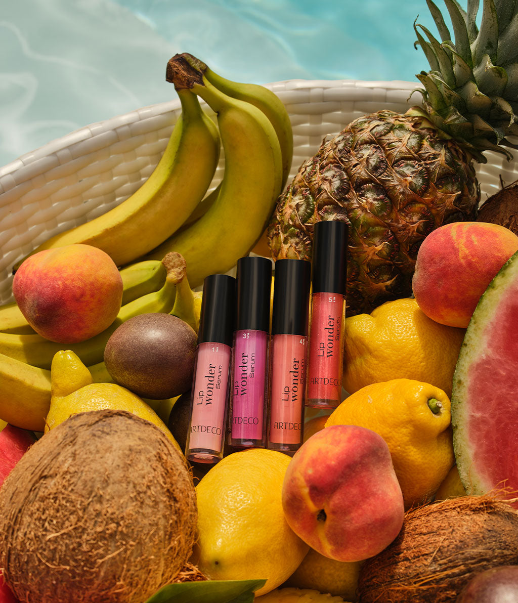 Lip Wonder Serum in vier sommerlichen Farben, platziert in einem Korb voller buntem Obst.