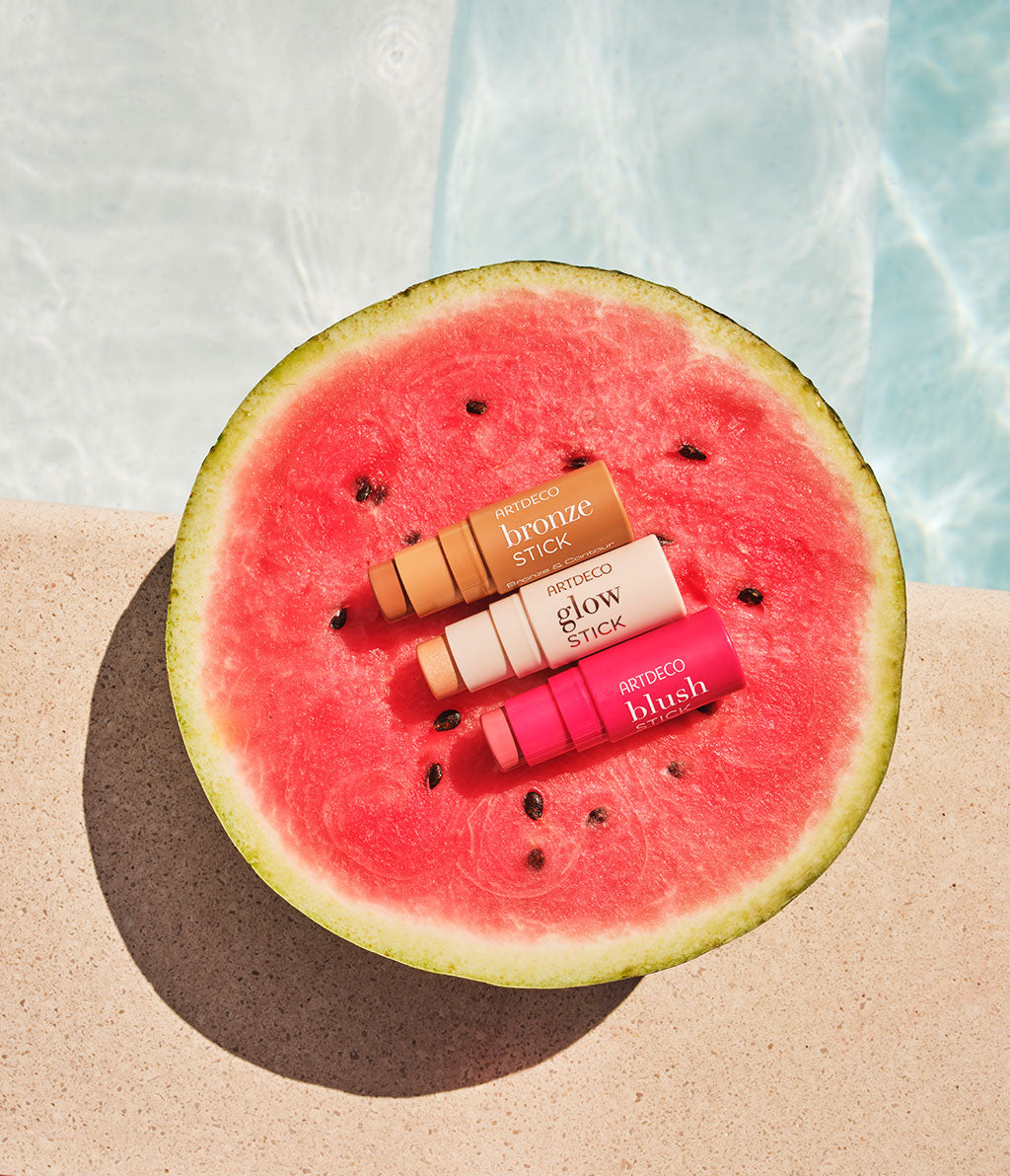 Bronze Stick, Blush Stick und Glow Stick liegen platziert auf einer halbierten Melone an einem Pool.