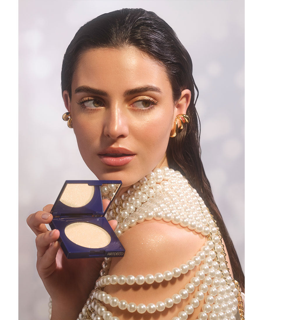 Brünettes Model blickt über ihre Schulter, hält das halbgeöffnete Diamond Beauty Powder in ihrer Hand. Der champagnerfarbene All-over-Highlight würde dabei in ihrem Gesicht als auch auf den Schultern, für einen unwiderstehlichen Glow aufgetragen