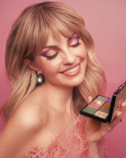 Blondes Model hält die limitierte Eyes Eyes Baby Palette mit acht metallischen und Pearl-Tönen, ideal für kreative Augen-Make-ups, vor einem rosa Hintergrund.