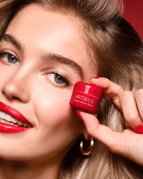 Junge Frau mit blonden Haaren hält die Eyeshadow Base im limitierten roten Design in der Hand und lächelt.