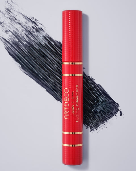 Lash Hero Tubing Mascara im limitierten roten Design liegt geschlossen auf einem neutralen Hintergrund. Zusätzlich ist ein Textur-Swatch der Mascara auf dem Hintergrund.