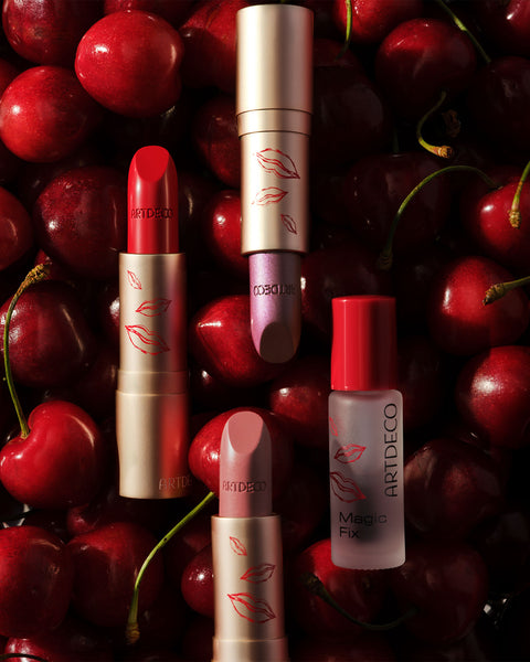Drei geöffnete Perfect Color Lipsticks liegen zusammen mit dem Magic Fix im limitierten roten Design auf einem Untergrund aus Kirschen.