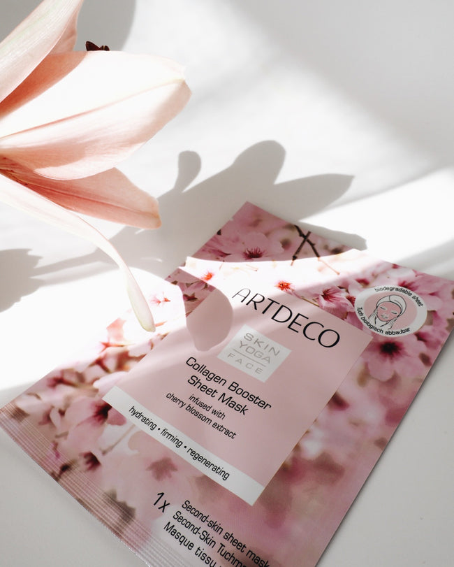 Collagen Booster Maske liegt auf einem weißen Hintergrund und eine Blume wird davorgehalten.