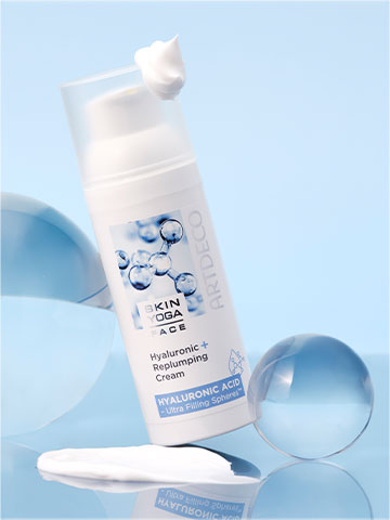 Die Hyaluronic + Replumping Cream vor einem hellblauen Hintegrund umgeben von verschieden großen Glaskugeln. Auf dem Deckel ist etwas Creme-Klecks zu sehen.
