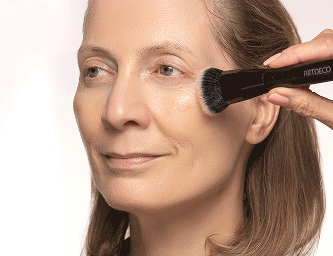 Eine feuchtigkeitsspendende Foundation wird mit einem Pinsel aufgetragen und mögliche Unebenheiten mit einem Concealer kaschiert