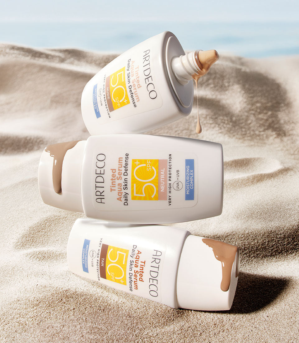 Tinted Aqua Serum Daily Defense SPF50+ liegt im Sand und die einzelnen Farben sind aufeinander gestapelt