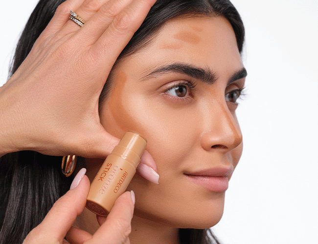 Model legt ihre Hand in L-Form an ihre Wangenknochen, um die perfekte Bronzer Platzierung zu erhalten. Im nächsten Schritt wird Bronzer am Stirnansatz aufgetragen und anschließend verblendet.