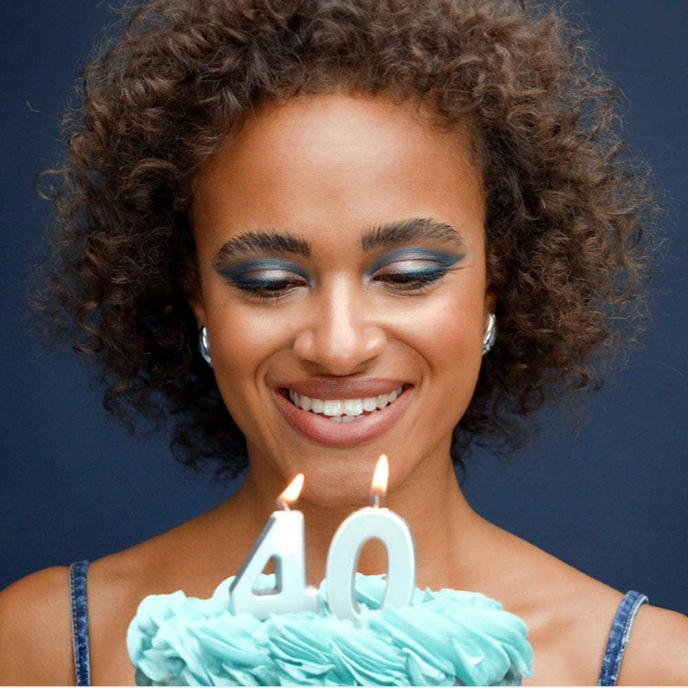 Frau mit braunem lockigem Haar und blauem Augen Make-Up hält eine Torte vor sich. Auf der Torte sind Kerzen, die die Zahl 40 zeigen. 