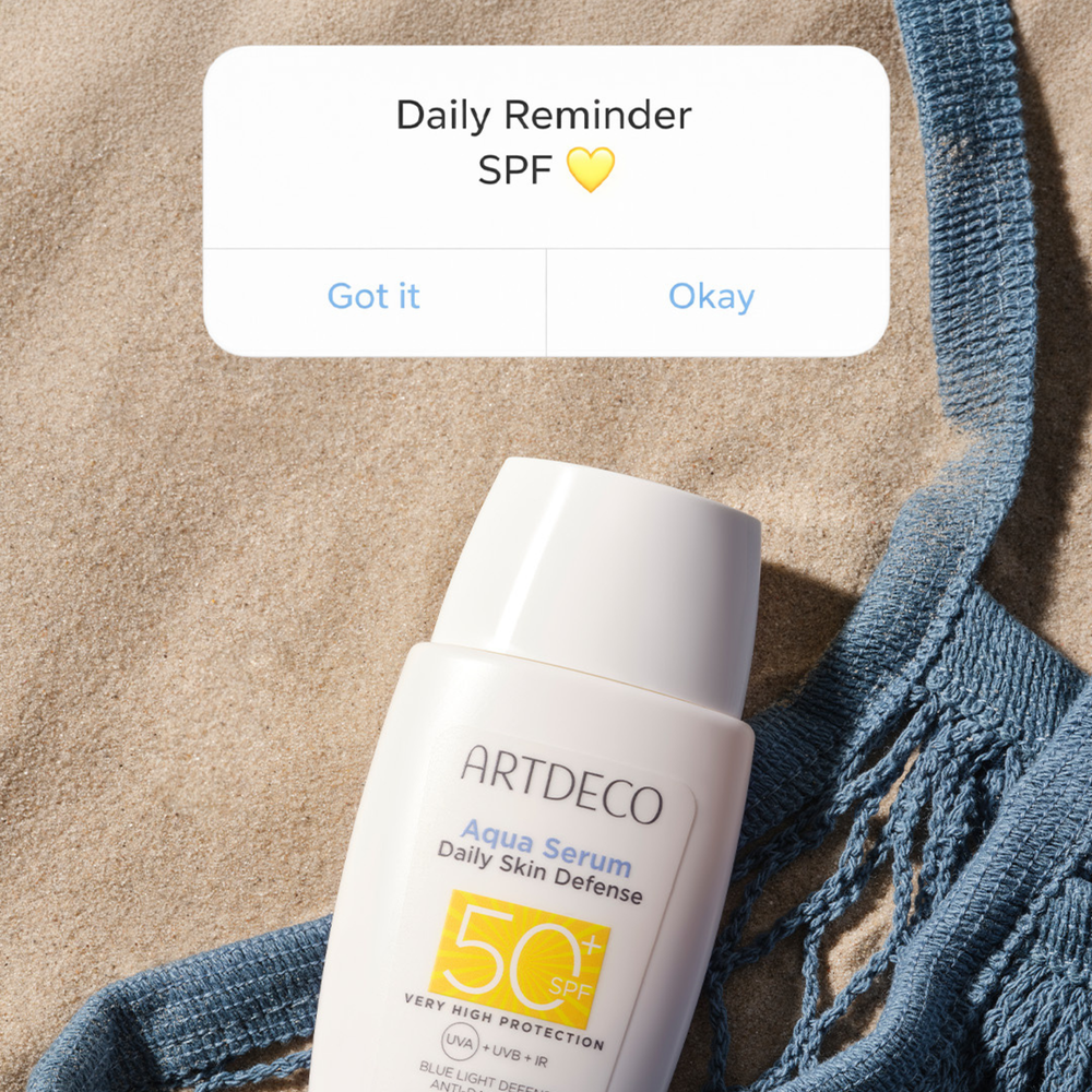 Aqua Serum Daily Defense SPF50+  liegt im Sand. Darüber wird grafisch ein  Reminder zum Sonnenschutz angezeigt