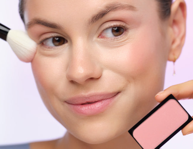 Model trägt mit einem Puderpinsel den pinken Blush auf, welchen sie in ihrer anderen Hand hält