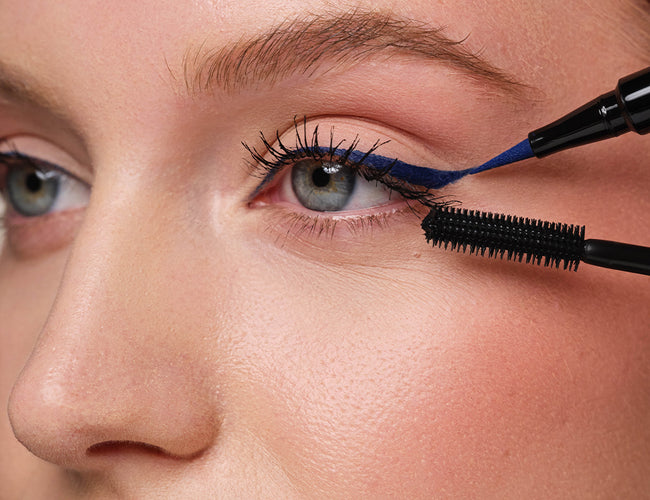 Close-up auf das Auge des Models, auf welchem ein blauer Eyeliner aufgetragen und zusätzlich eine schwarze Mascara appliziert, wird