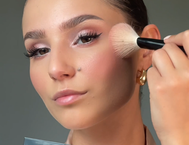 Model trägt einen Blush mit einem fluffigen Pinsel auf die Wangen auf.