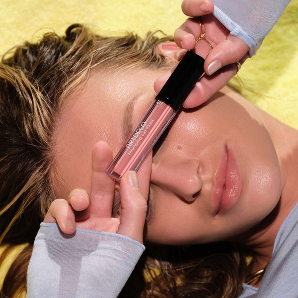 Model liegt auf einem gelben Handtuch und hält einen ARTDECO Plumping Lip Fluid Lipgloss in Rosé vor ihr Gesicht.