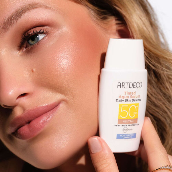 Nahaufnahme eines Models mit strahlender Haut, die das ARTDECO Tinted Aqua Serum Daily Skin Defense SPF 50+ neben ihrem Gesicht hält.